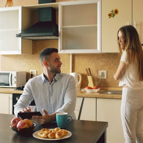 Happy young man using digital tablet computer sitting in the kitchen and talking his wife while she cooking at home in the morning