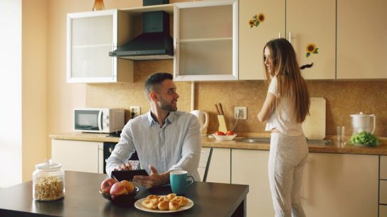 Happy young man using digital tablet computer sitting in the kitchen and talking his wife while she cooking at home in the morning